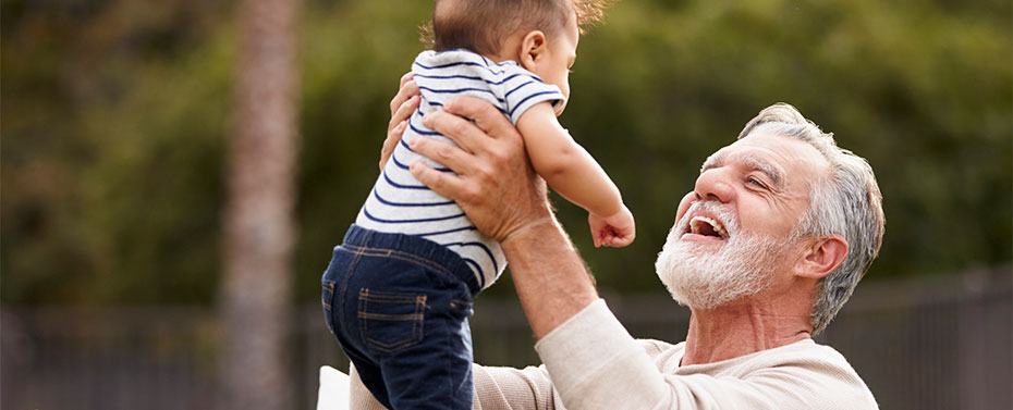 Man sitting in the garden lifting his baby grandson in the air and smiling to him